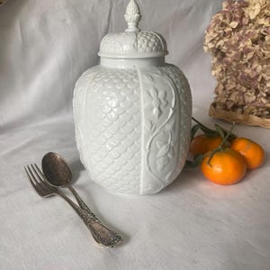 Puede incluir: Un jarrón de cerámica blanco con tapa, con un patrón texturizado en forma de escamas y detalles de relieve floral. El jarrón está acompañado de un tenedor y una cuchara antiguos, y unas pequeñas naranjas. El jarrón mide unos 25 cm de alto.