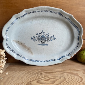 Peut inclure: Plat ovale de service de style ancien, avec un bord festonné. La plat en céramique blanche présente un motif floral bleu au centre et une bordure bleue à motifs. Le plat montre des signes d'âge, ajoutant à son charme vintage. Une pièce décorative pour servir ou exposer.