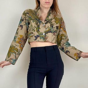 Può includere: Una giacca corta con bottoni e motivo floreale nei toni del verde, giallo e blu. Abbinata a pantaloni neri a vita alta e svasati. L'outfit è un insieme alla moda.