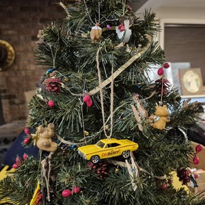 Puede incluir: Un árbol de Navidad decorado con diversos adornos, incluyendo un coche de juguete amarillo con texto "Oh Henry!", pequeñas figuras de animales y racimos de bayas rojas. El árbol está situado contra una rústica pared de ladrillo.