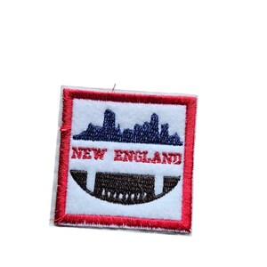 Puede incluir: Un parche blanco bordado con un borde rojo. El parche presenta un balón de fútbol blanco y negro con una silueta azul de un horizonte de ciudad por encima. El texto "NEW ENGLAND" está escrito en rojo sobre el balón.