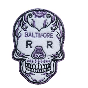 May include: Embroidered patch featuring a sugar skull design. The skull is white with purple accents and decorative floral elements. The text "BALTIMORE" and two "R" letters are visible. The patch has a black border.