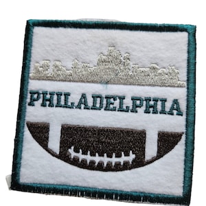 May include: A white embroidered patch with a teal border. The patch features a silver embroidered skyline of Philadelphia, the text "PHILADELPHIA" in teal, and a brown football.