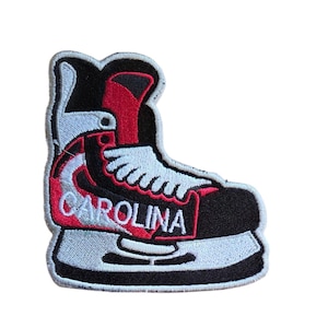 May include: An embroidered patch shaped like an ice hockey skate in black, white, and red. The skate features white laces, a white blade holder, and the word CAROLINA stitched in white capital letters across the side of the boot section.