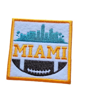 May include: An orange embroidered patch with a white background. The patch features a blue silhouette of the Miami skyline and the word "MIAMI" in yellow letters. A black and white football is below the skyline.