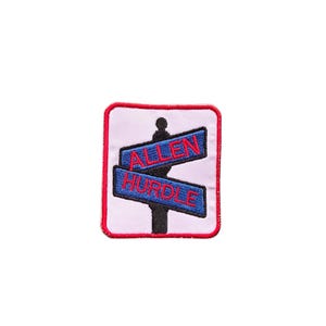 May include: Embroidered patch featuring a signpost design. The signposts read "ALLEN" and "HURDLE" in red lettering on blue backgrounds. The patch has a white background with a red border. The signpost is black.