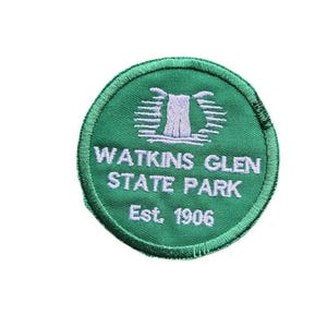 May include: A circular green embroidered patch featuring a white graphic of a waterfall. The text reads WATKINS GLEN STATE PARK and Est. 1906 in white thread. The patch has a stitched border and is designed for attachment to fabric items.