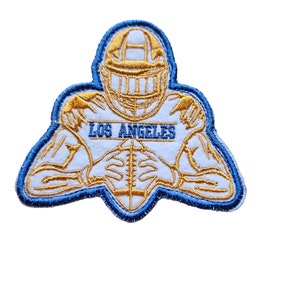 Peut inclure: Un écusson brodé bleu et or représentant un joueur de football américain portant un casque avec le texte "LOS ANGELES" sur la poitrine.