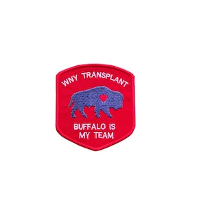 May include: A red embroidered patch with the text "WNY TRANSPLANT" and "BUFFALO IS MY TEAM". The patch features a blue buffalo silhouette with a red heart. The patch is shield-shaped.