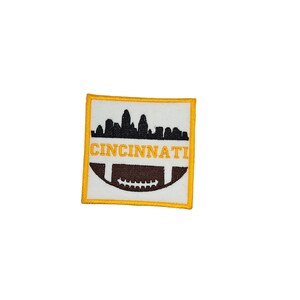 May include: A yellow and brown embroidered patch with a silhouette of the Cincinnati skyline and a football. The text "CINCINNATI" is embroidered in yellow.