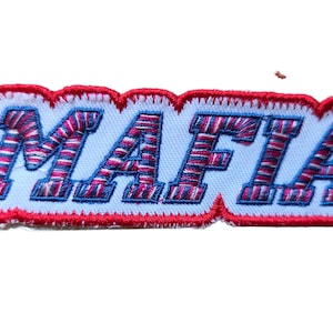 Op de afbeelding: Geborduurde patch met het woord "MAFIA" in hoofdletters. De letters zijn wit met rode en blauwe stiksels, omlijnd in blauw en omrand door een rode zigzagsteek op een witte achtergrond.