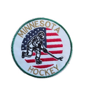May include: A circular embroidered patch featuring a hockey player in black thread over a background of the American flag. The text MINNESOTA is arched in gold at the top, and HOCKEY is arched in gold at the bottom, all bordered by green.