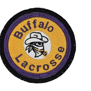 May include: A round, embroidered patch with a purple and black border. The centre is yellow with the words "Buffalo Lacrosse" in purple lettering. A white cartoon image of a person wearing a cowboy hat and bandana is in the centre.