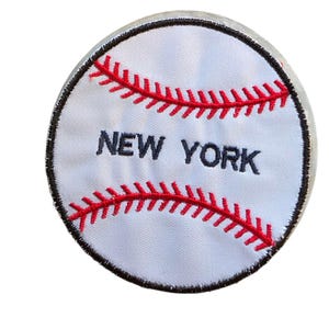 May include: A circular embroidered patch featuring a white baseball design with red stitching details and the text NEW YORK in dark blue lettering. The patch has a thick black border around the entire circumference of the circular shape.