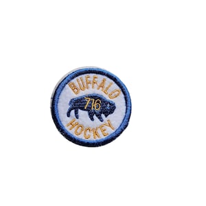 May include: A white, round patch with a blue border. The patch features a gold embroidered buffalo with the text "BUFFALO 716 HOCKEY" around the buffalo.