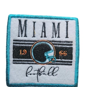 May include: A white embroidered patch with a teal border. The patch features the word "MIAMI" in black lettering, a black and white football helmet with a teal facemask, and the numbers "19" and "66" in orange. The word "football" is written in cursive below the helmet.