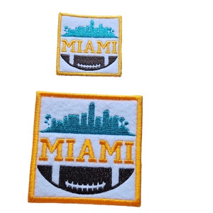 May include: Two embroidered patches with a white background, a gold outline, and a black and white football graphic. The top patch is smaller and features a teal skyline with the word "MIAMI" in yellow. The bottom patch is larger and features a teal skyline with the word "MIAMI" in yellow.