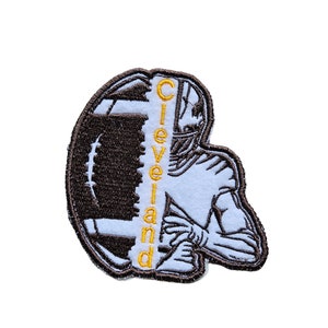 May include: An embroidered patch featuring a football player in a white uniform with a brown football. The word "Cleveland" is embroidered vertically in yellow on the left side of the patch.
