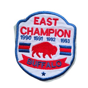 May include: Embroidered patch with a white background, blue border, and red accents. The patch features the text "EAST CHAMPION" above the years 1990-1993. A red buffalo and the word "BUFFALO" are also displayed.