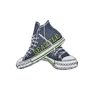 Puede incluir: Parche bordado de un par de zapatillas azules con cordones blancos y un contorno verde. La palabra "Seattle" está bordada en verde en el lateral de las zapatillas.