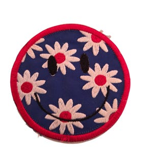 May include: Round patch with a smiley face design. The face is navy blue with white daisies and a red border. The smiley face has black eyes and a black curved mouth.