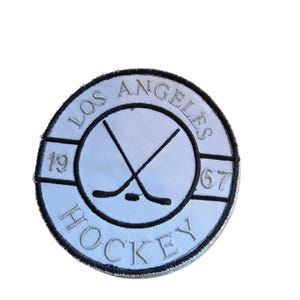 Könnte beinhalten: Ein kreisförmiger gestickter Aufnäher mit hellblauem Hintergrund und schwarzem Rand. In der Mitte sind zwei gekreuzte schwarze Hockeyschläger abgebildet. Der Text LOS ANGELES steht oben, HOCKEY unten, und 19 sowie 67 an den Seiten.