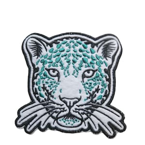 Op de afbeelding: Een geborduurde patch van een wit sneeuwluipaard gezicht met turquoise vlekken en zwarte snorharen.