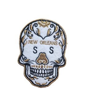 May include: White skull patch with gold and black embroidery. The design includes floral accents, the words "NEW ORLEANS", and the letters "S S". The skull has a gold nose and a detailed tooth design.