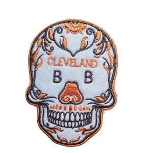 May include: An embroidered patch featuring a white skull with orange and black details. The skull has a floral design around the eyes and mouth. The text "CLEVELAND B B" is embroidered in orange on the skull's forehead.