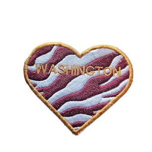 May include: A heart-shaped patch with a white and burgundy zebra stripe pattern. The patch is outlined in gold thread and has the word "WASHINGTON" embroidered in gold thread.