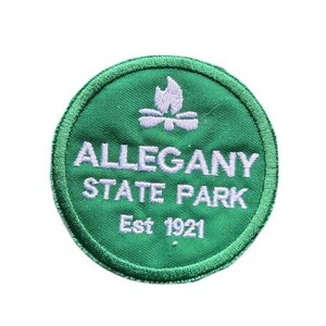 May include: A circular green embroidered patch featuring a white campfire icon at the top. The text reads ALLEGANY STATE PARK in white capital letters, with Est 1921 printed below in a smaller white font. The patch has a stitched green border.