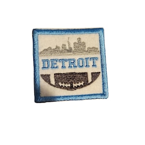 May include: An embroidered patch with a blue border, featuring a white background with a gray cityscape silhouette and the word "DETROIT" in black text. A brown football is depicted below the city skyline.