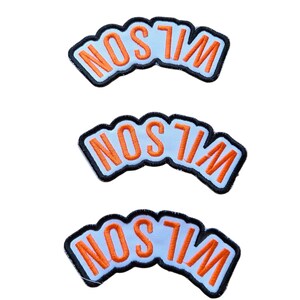 May include: Three embroidered patches with the name "WILSON" in orange letters on a light blue background. Each patch has a black border and a curved shape, suitable for application on clothing or accessories.