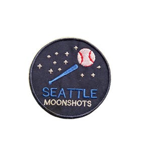 May include: A navy blue circular patch featuring a baseball bat and ball, with stars and the text "SEATTLE MOONSHOTS" in white and blue. The baseball is red and white. This patch is likely for a sports fan.
