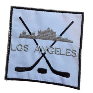 May include: A square embroidered patch with a white background and a black border. It features two crossed black hockey sticks, a black hockey puck, a grey skyline silhouette, and the grey embroidered text LOS ANGELES centered on the patch.