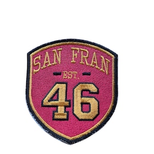 Puede incluir: Un parche en forma de escudo con las palabras "SAN FRAN" bordadas en oro, encima del número "46". El parche tiene un fondo rosa oscuro con un borde dorado y el texto "EST." en oro.