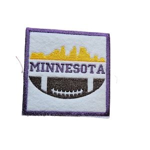 Op de afbeelding: Paarse en witte geborduurde patch met een geel silhouet van de skyline van Minneapolis, het woord "MINNESOTA" en een bruine voetbal.