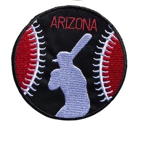 May include: A circular embroidered patch featuring a black background with a red baseball design on the sides, white stitching details, a light blue silhouette of a baseball player holding a bat in the center, and the red text ARIZONA above.
