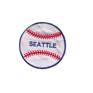 May include: A white baseball patch with red stitching and the word "SEATTLE" in blue lettering. The patch has a blue border and is designed to resemble a baseball.