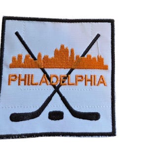 May include: A square embroidered patch with a white background and a black border. It features an orange skyline silhouette of Philadelphia above the word PHILADELPHIA in orange text, with two crossed black hockey sticks and a black puck below.