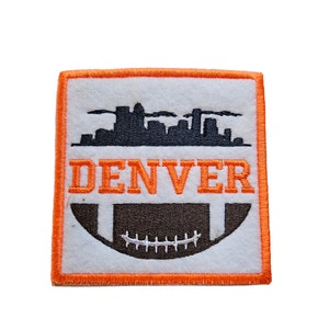 Könnte beinhalten: Orangefarbener und weißer gestickter Aufnäher mit einer schwarzen Silhouette der Skyline von Denver und einem braunen Fußball mit weißen Schnürsenkeln. Der Text "DENVER" ist in Schwarz gestickt.