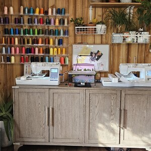 May include: Two white sewing machines with colorful spools of thread on shelves behind them. The machines are on a wooden cabinet with three doors.