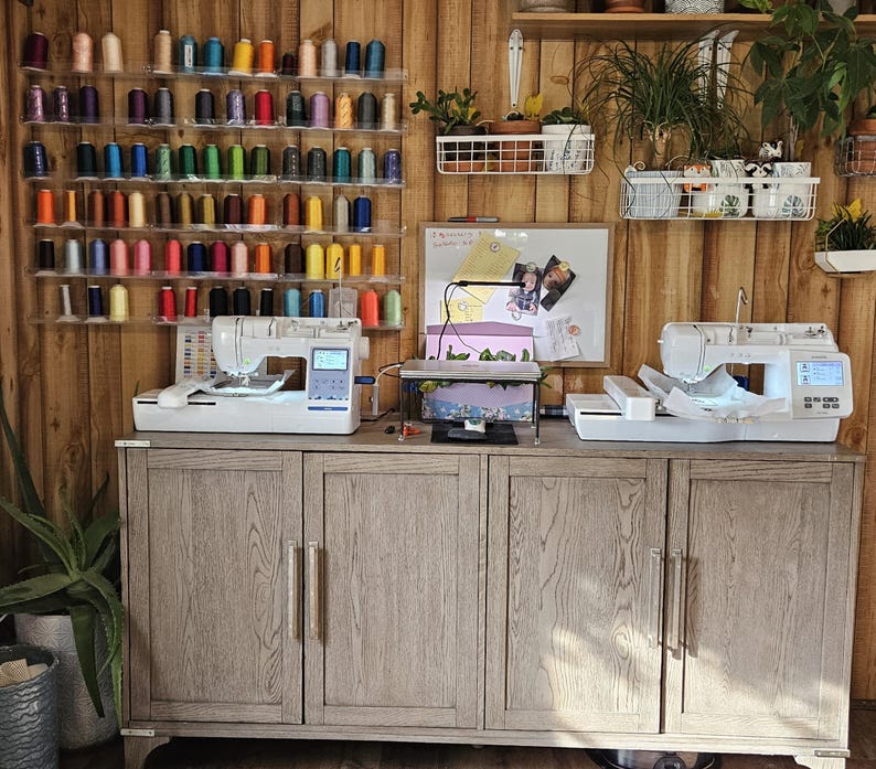 May include: Two white sewing machines with colorful spools of thread on shelves behind them. The machines are on a wooden cabinet with three doors.