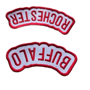 May include: Two embroidered patches with white fabric and red stitching. The top patch reads "ROCHESTER" in red letters. The bottom patch reads "BUFFALO" in red letters.