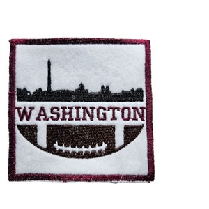 May include: A burgundy embroidered patch with a white background. The patch features a black silhouette of the Washington D.C. skyline with the Washington Monument in the centre. The text "WASHINGTON" is embroidered in black above a brown football.