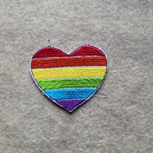 May include: A rainbow heart-shaped patch with red, orange, yellow, green, blue, and purple stripes. The patch is embroidered and has a white outline.