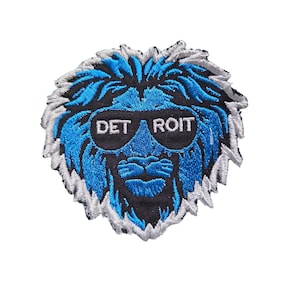 May include: Embroidered patch featuring a blue lion's head wearing sunglasses. The sunglasses have the text "DETROIT" on them. The lion has a blue face, black nose and mouth, and a white and gray mane. The patch is on a white background.