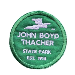 May include: A circular green embroidered patch featuring white stitched text that reads JOHN BOYD THACHER STATE PARK EST. 1914. Above the text is a stylized white graphic of a bird in flight. The patch has a thick, stitched green border.