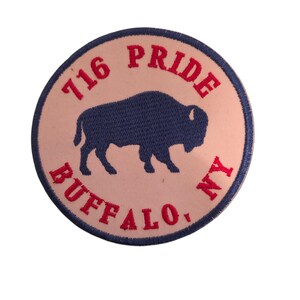 Może przedstawiać: Okrągła, haftowana naszywka z granatową sylwetką bizona na jasnoróżowym tle. Napis "716 PRIDE BUFFALO, NY" jest wyhaftowany na czerwono wokół krawędzi naszywki.