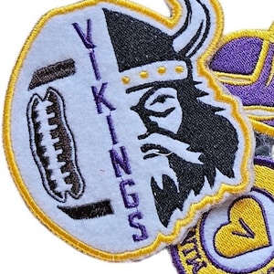 Pode incluir: Patch bordado com um viking com capacete com chifres e a palavra "Vikings" em roxo. O patch tem um fundo branco com um design de futebol americano e uma borda dourada. Patches adicionais são visíveis.